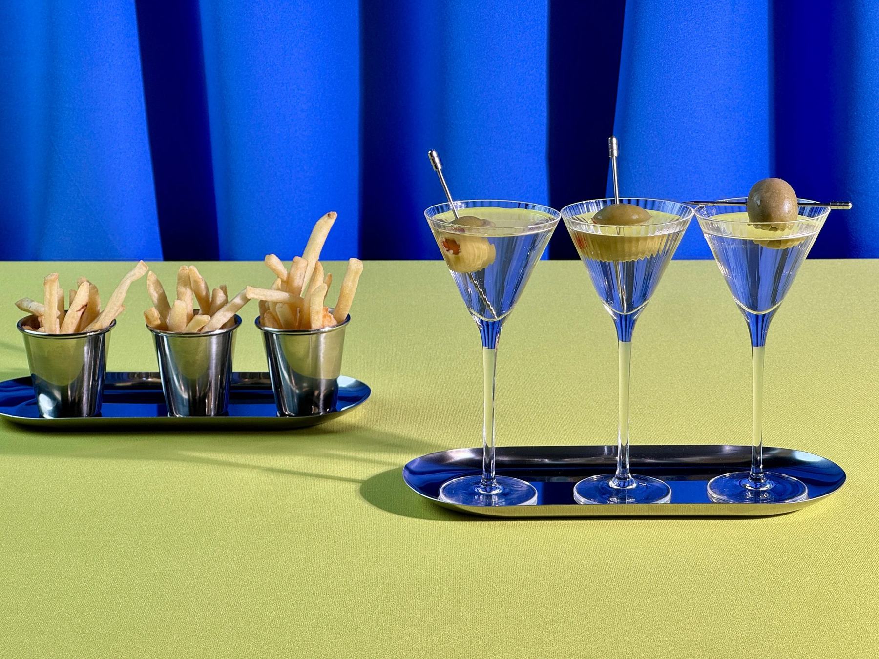 Three mini martinis with olives on a silver tray beside cups of fries, against a blue curtain